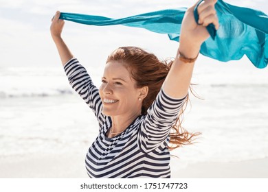 Smiling Mature Woman Holding Blue Scarf Over Head While Walking At Beach. Happy Active And Healthy Woman Enjoying Summer Vacation At Sea. Middle Aged Lady Running At Seashore With Blue Fabric.