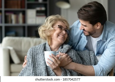 Smiling mature woman holding cup, hugging with adult son close up, enjoying leisure time, happy elderly mother wearing warm scarf drinking hot tea or coffee, family spending weekend together at home - Powered by Shutterstock