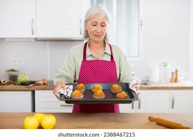 Smiling mature woman holding tray with freshly baked muffins in home kitchen - Powered by Shutterstock