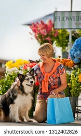 Smiling Mature Woman Kneeling Down To Pet Her Dog At A Flower Market.