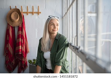 Smiling mature woman looking outside window. Thoughtful old woman looking away through window. Senior beautiful woman sitting at home with pensive expression.
