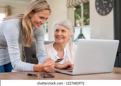 Smiling Mature Woman Making Payment Via Credit Card For Shopping Done By Senior Woman At Home. Elderly Mother And Daughter Buying Online. Family Generations Using Laptop Computer Paying Bills.