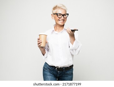 Smiling Mature Woman With Short Gray Hair Holding Mobile Phone And Cup Of Takeaway Coffee Over Grey Background. Lifestyle, Business And Old People Concept.