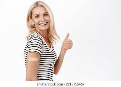 Smiling Mature Woman, Shows Thumbs Up, Has Covid-19 Vaccination Band Aid On Shoulder, Recommends People Coronavirus Vaccine, White Background