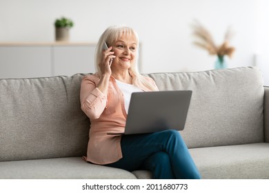 Smiling Mature Woman Talking On Phone Using Laptop Working Distantly Sitting On Couch At Home. Modern Middle-Aged Lady Communicating Speaking Via Cellphone. Selective Focus. Elderly And Technology