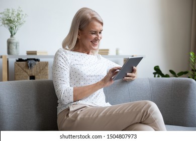 Smiling Mature Woman Using Computer Tablet At Home, Happy Older Female Sitting On Couch, Holding Electronic Device, Looking At Screen, Reading News In Social Network, Shopping Or Chatting Online