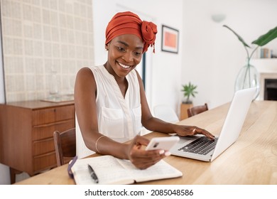 Smiling Mature Woman Using Smartphone While Working On Laptop From Home. African Middle Aged Businesswoman Checking Notification On Smart Phone While Taking A Break During Work. Smart Working.