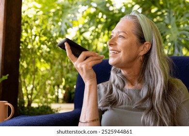 Smiling mature woman using smartphone voice assistant while relaxing on chair at home. Technology, relaxation, lifestyle, communication, leisure, comfort - Powered by Shutterstock