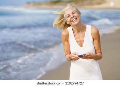 Smiling mature woman walking on the beach using a smartphone. - Powered by Shutterstock