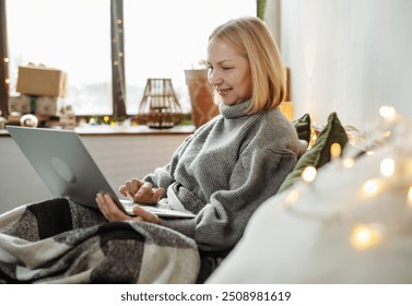 Smiling mature woman watching movie on laptop on bed under blanket at home. Magic of the holiday. Christmas time. Use technology - Powered by Shutterstock