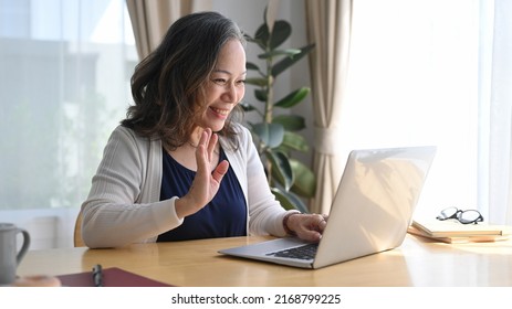 Smiling mature woman waving hand during video conference while remote working from home - Powered by Shutterstock