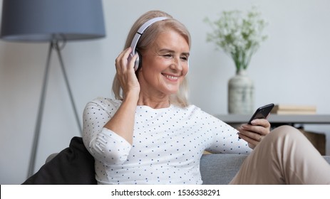 Smiling Mature Woman Wear Wireless Headphone Hold Smartphone Looking At Phone Screen Using Mobile Player App Listening Online Music, Learning Foreign Language, Watching Video Relaxing On Sofa At Home