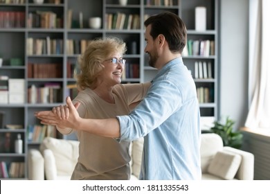 Smiling Mature Woman Wearing Glasses Dancing With Adult Son In Modern Living Room At Home, Young Man And Happy Older Mother Enjoying Leisure Time Together, Moving To Music, Two Generations