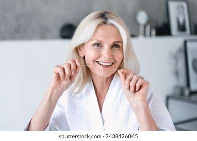 Smiling mature woman in white bathrobe standing in domestic bathroom and clean teeth using dental floss. Happy female doing morning hygiene procedure. Oral care concept - Powered by Shutterstock