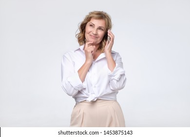 Smiling Mature Woman In White Shirt Talking On The Phone With Her Business Partner. Studio Shot. Positive Facial Emotion.