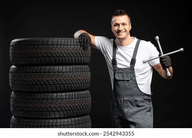 Smiling mechanic holding lug wrench and leaning on a stack of new winter tires on black background - Powered by Shutterstock