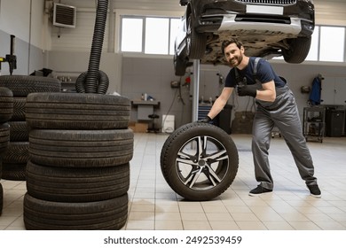 Smiling mechanic in overalls and gloves working in auto repair shop. Mechanic handling tire in workshop with car lifted in background. Professional automotive maintenance and tire service. - Powered by Shutterstock