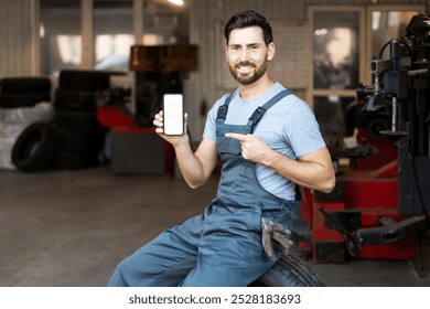 Smiling mechanic sitting on tire and pointing at smartphone with blank screen in auto repair shop - Powered by Shutterstock