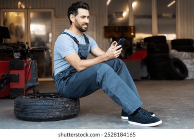 Smiling mechanic sitting on a tire browsing on his mobile phone in a auto repair shop - Powered by Shutterstock