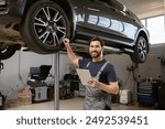 Smiling mechanic using tablet in auto repair shop with car on lift. Confident male technician diagnosing vehicle problem using modern technology. Professional car service in well-equipped garage.