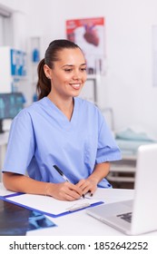 Smiling Medical Nurse Writing Notes On Clipboard Sitting At Desk In Hospital. Health Care Physician Using Computer In Modern Clinic Looking At Monitor, Medicine, Profession, Scrubs.