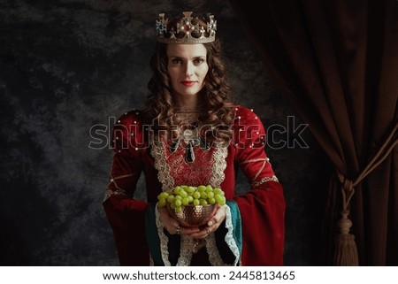 smiling medieval queen in red dress with plate of grapes and crown on dark gray background.