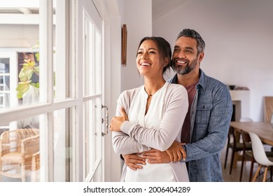 Smiling mid adult couple hugging each other and standing near window while looking outside. Happy and romantic mature man embracing hispanic wife from behind while standing at home with copy space.