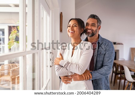 Smiling mid adult couple hugging each other and standing near window while looking outside. Happy and romantic mature man embracing hispanic wife from behind while standing at home with copy space.