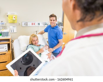 Smiling Mid Adult Couple With Newborn Babygirl Looking At Doctor Holding Digital Tablet In Hospital