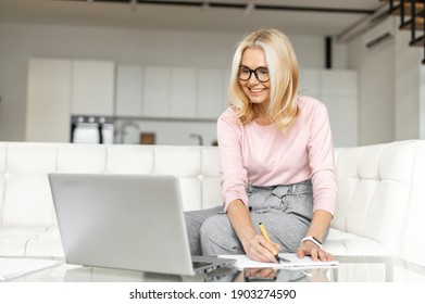 Smiling mid age female office worker business lady listening to an online webinar on laptop, taking notes in notebook, working on freelance project at home, remote work, learning at home concept