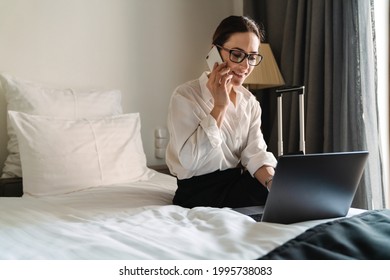 Smiling Mid Aged Brunette White Businesswoman Sitting On Bed In A Hotel Room Talking On Mobile Phone Using Laptop Computer