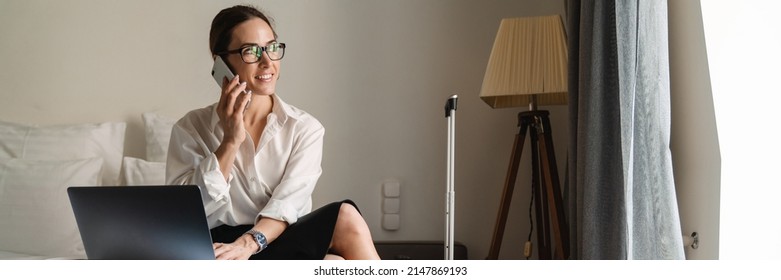 Smiling Mid Aged Brunette White Businesswoman Sitting On Bed In A Hotel Room Talking On Mobile Phone Using Laptop Computer