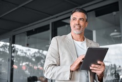 Smiling Mid Aged Business Man Wearing Suit Standing Outside Office Holding Digital Tablet. Mature Businessman Professional Holding Fintech Device Looking Away Thinking Or New Business Ideas Solutions.