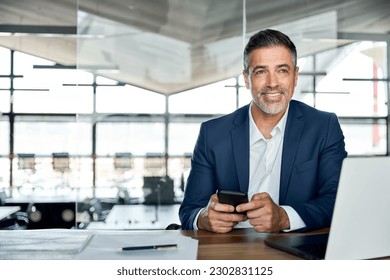 Smiling mid aged business man ceo wearing blue suit sitting in office using cell phone solutions. Mature businessman professional executive holding mobile working at desk with laptop and smartphone.