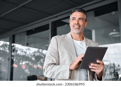 Smiling mid aged business man wearing suit standing outside office holding digital tablet. Mature businessman professional holding fintech device looking away thinking or new business ideas solutions.