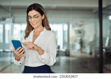 Smiling mid aged business woman holding cell phone standing in office. Happy mature professional businesswoman wearing eyeglasses holding smartphone using smartphone looking at cellphone, copy space.