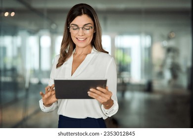 Smiling mid aged mature professional business woman bank manager, 40s female executive, entrepreneur holding fintech tab digital tablet corporate digital technology device standing in office at work. - Powered by Shutterstock