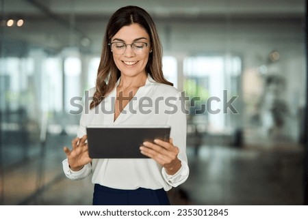 Smiling mid aged mature professional business woman bank manager, 40s female executive, entrepreneur holding fintech tab digital tablet corporate digital technology device standing in office at work.