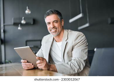 Smiling mid aged successful business man using digital tablet computer sitting in modern office. Happy mature older professional businessman entrepreneur working on technology device, portrait. - Powered by Shutterstock