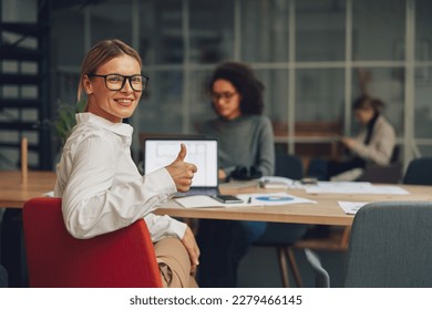 Smiling middle age businesswoman showing thumb up while working on laptop on colleagues background - Powered by Shutterstock