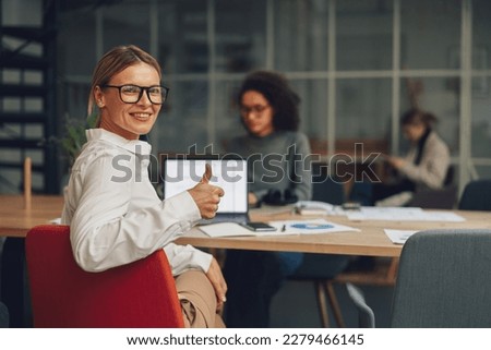 Smiling middle age businesswoman showing thumb up while working on laptop on colleagues background