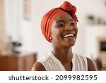 Smiling middle aged african american woman with orange headscarf. Beautiful black woman in casual clothing with traditional turban at home laughing. Portrait of mature carefree lady looking away.