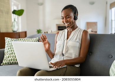 Smiling Middle Aged African American Woman With Headphone And Mic Waving Hand During Video Call On Laptop. Cheerful Black Mid Adult Woman Enjoying Conference On Laptop While Sitting On Couch At Home.