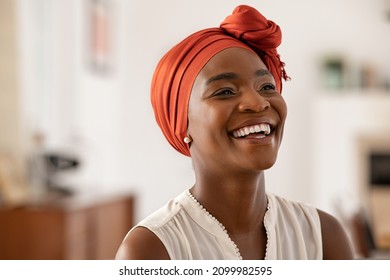 Lächelnde afrikanische Amerikanerin mittleren Alters mit orangefarbenem Kopftuch. Schöne schwarze Frau in zwangloser Kleidung mit traditionellen Turban zu Hause lachen. Portrait von reifer, sorgenfreier Dame, die wegschaut.