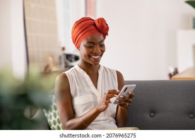 Smiling middle aged african woman with traditional head turban sitting on couch at home using smartphone. Beautiful african american woman with typical headscarf scrolling through internet on phone.