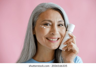 Smiling Middle Aged Asian Woman With Cream Drops Under Eye Holds Bottle Near Face On Pink Background In Studio. Mature Beauty Lifestyle