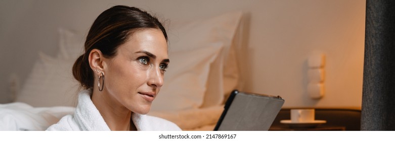 Smiling Middle Aged Brunette White Woman In Bathrobe Sitting On A Floor At The Bed Indoors Holding Tablet Computer