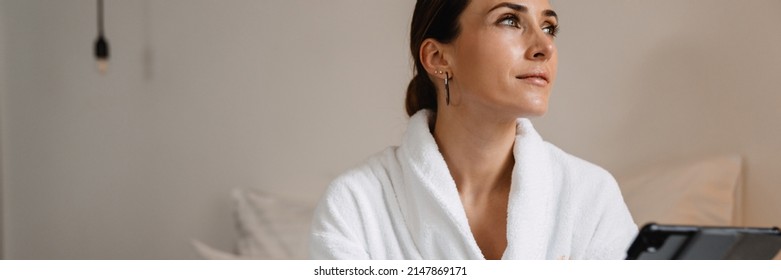 Smiling Middle Aged Brunette White Woman In Bathrobe Sitting On Bed Indoors Holding Tablet Computer