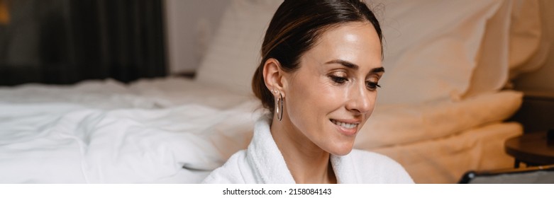 Smiling Middle Aged Brunette White Woman In Bathrobe Sitting On A Floor At The Bed Indoors Holding Tablet Computer