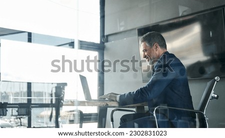 Smiling middle aged business man executive ceo sitting at desk using laptop. Happy professional businessman manager working on computer technology in office. View through glass.